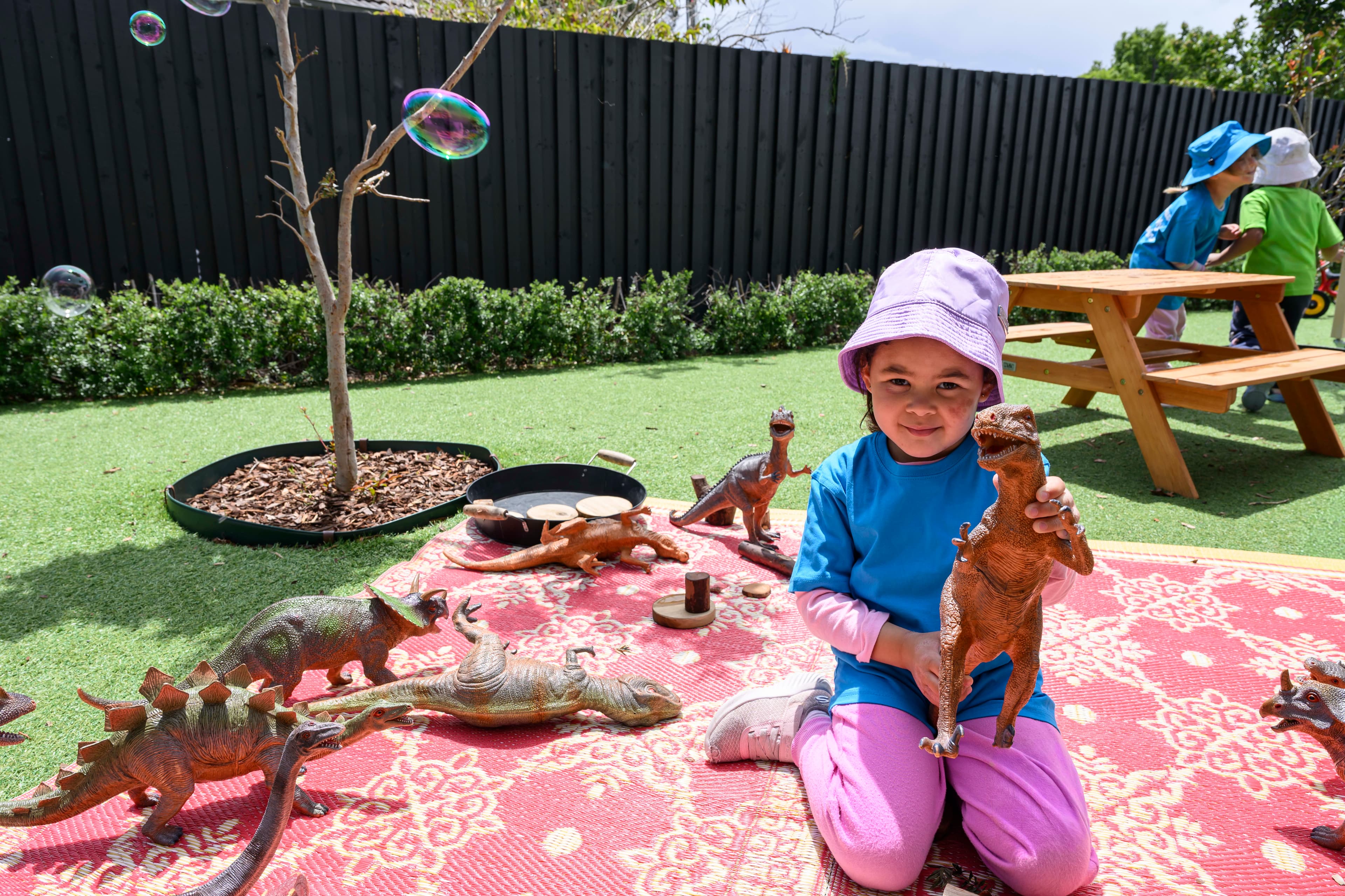 girl wearing blue shirt and pink pants playing with toy dinosaurs outdoors